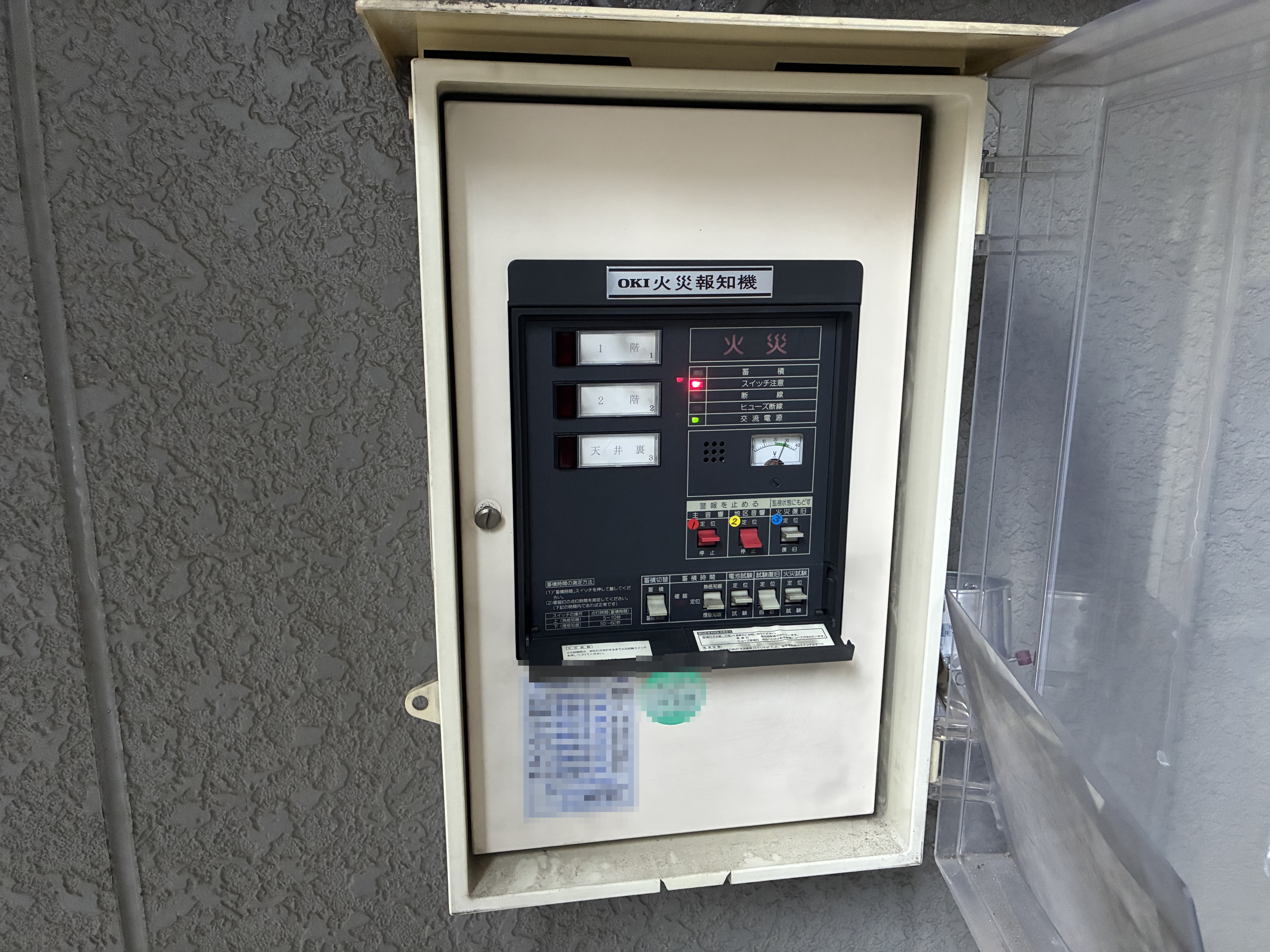 交換前の1993年製OKI火災受信機。1階・2階・天井裏の3回線表示とスイッチ注意ランプが点灯し、扉には当社の点検済証が貼られている（東京都板橋区）