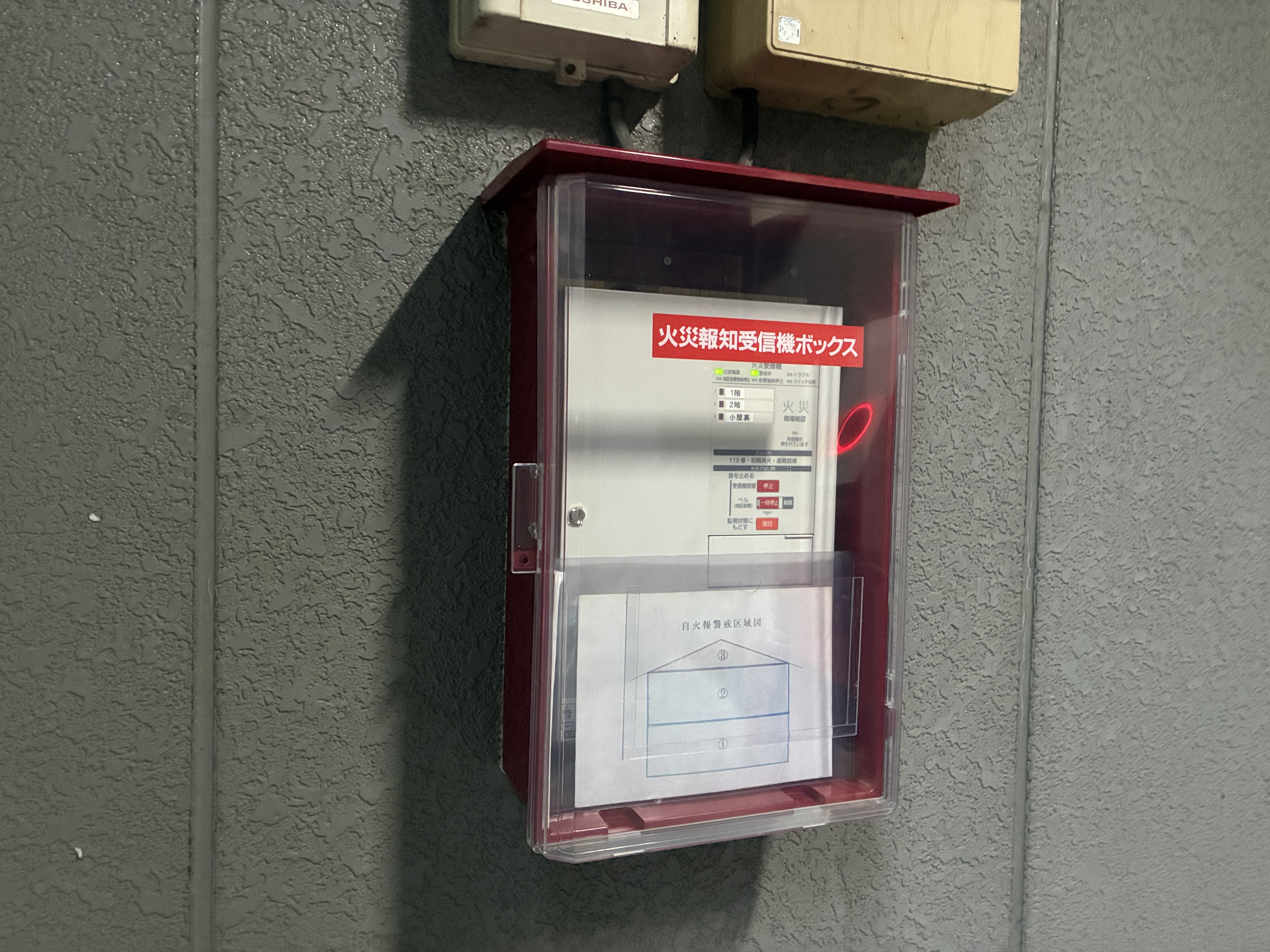 屋外に設置した火災報知受信機ボックス（透明扉のプラボックス）。内部に新しい受信機と自火報警戒区域図を収めている