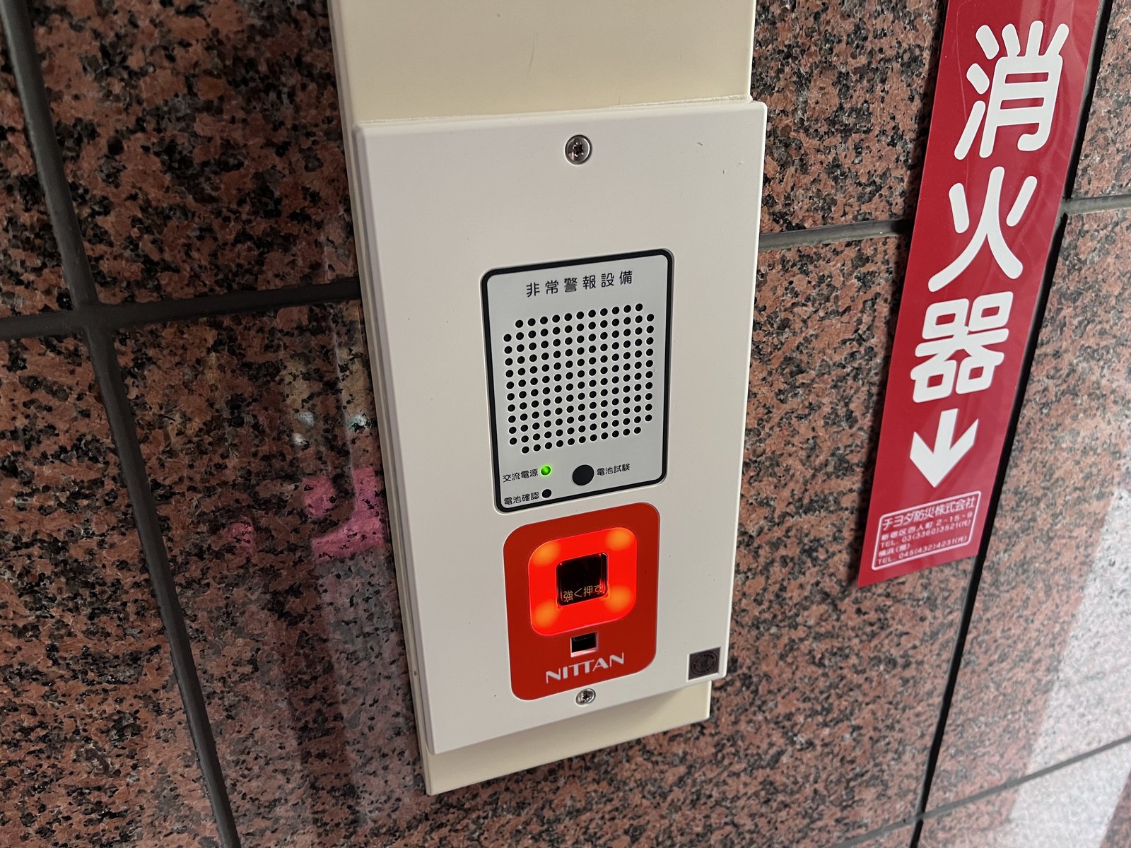交換後の新しい非常警報複合装置（ニッタン製）。上部にスピーカー、交流電源ランプ、電池試験ボタン、下部に赤い表示灯と押しボタン