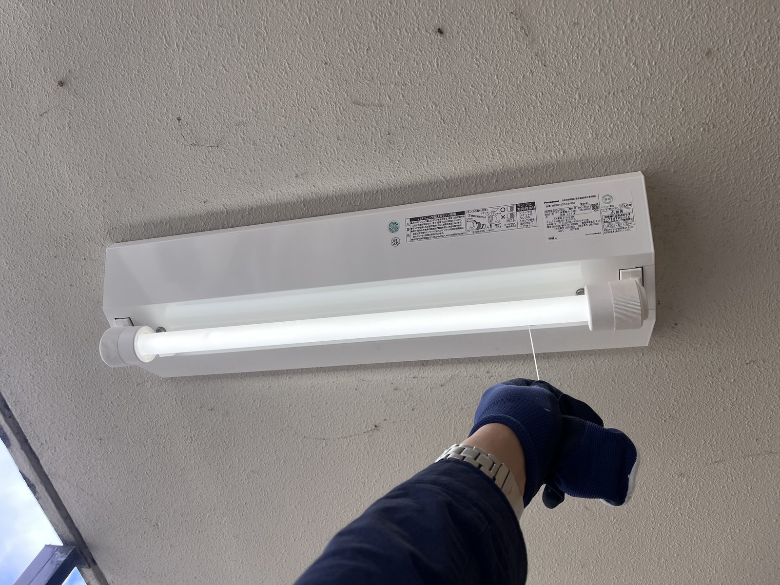 作業員が点検ひもを引いて、LED非常灯が予備電源（蓄電池）で点灯することを試験している様子