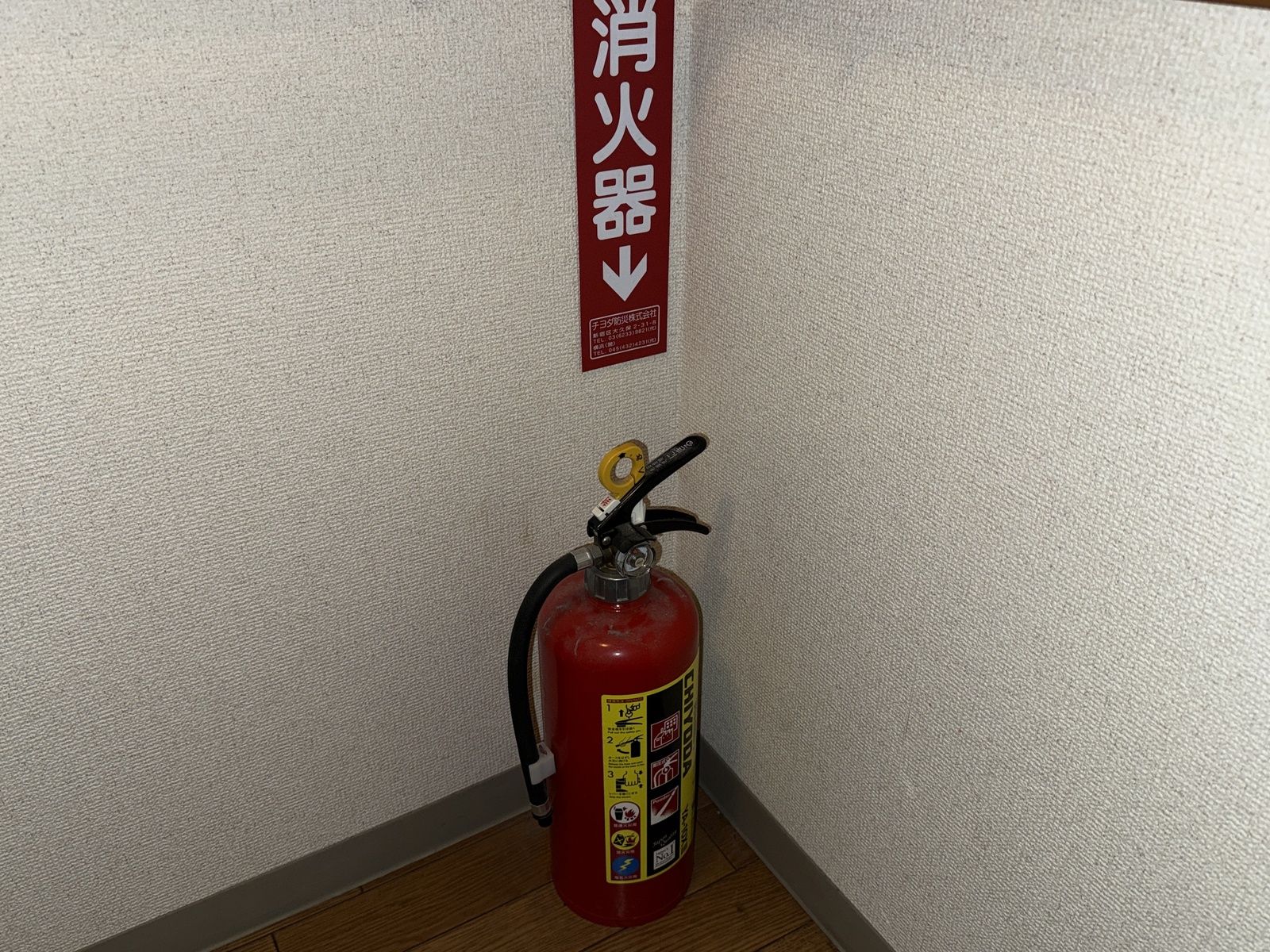 壁に「消火器」の標識が貼られた廊下の隅に置かれた交換前の粉末消火器