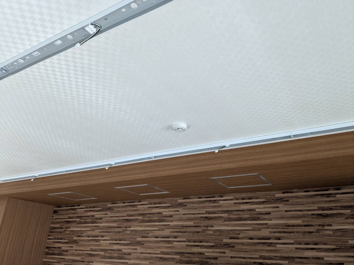 仕上げ後の天井に取り付けた光電式煙感知器。木目の下がり天井と白い天井クロスに合わせて納めている