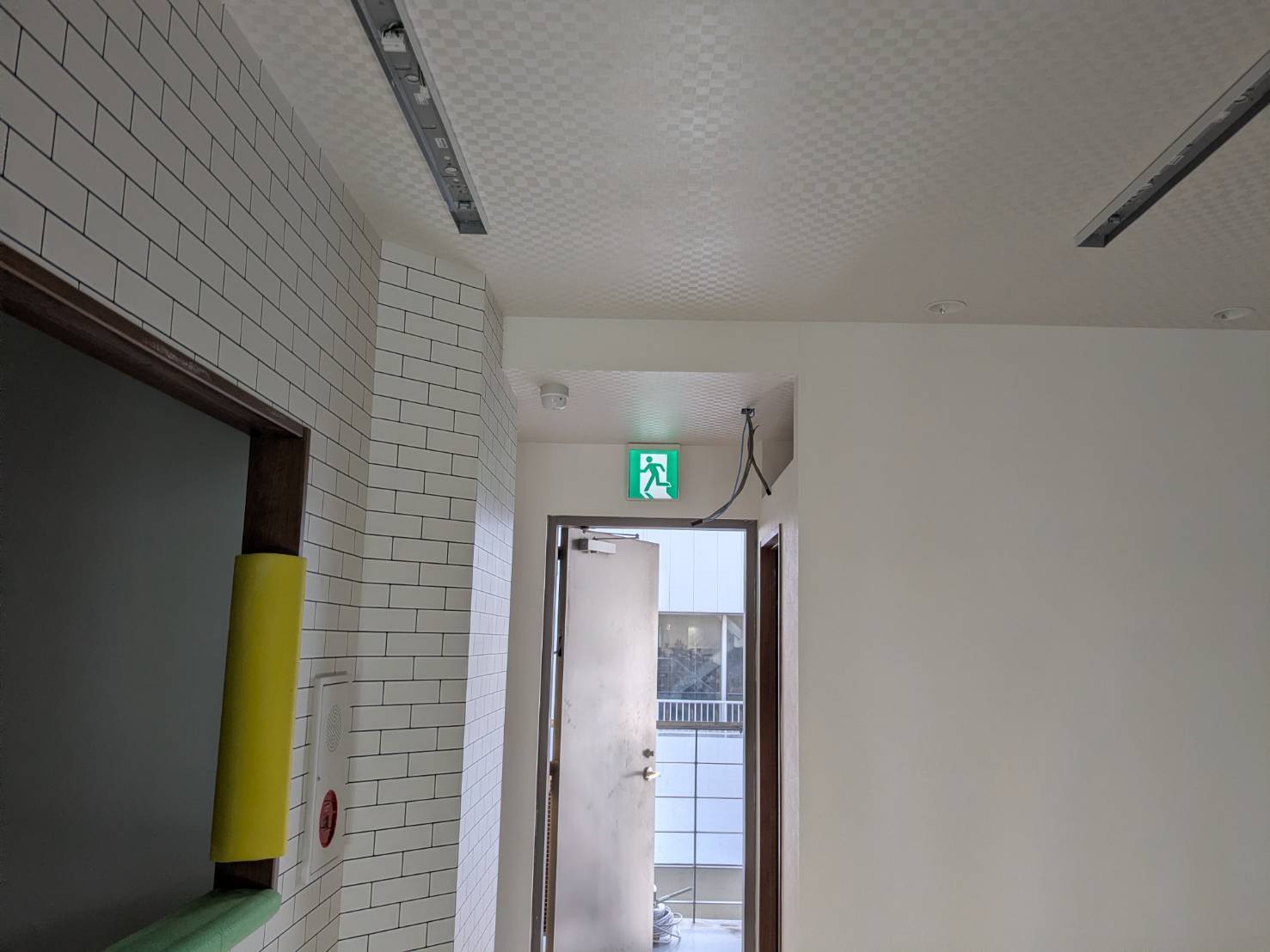 教室の出入口に取り付けた誘導灯と煙感知器。白いタイル壁の受付横に総合盤も見える