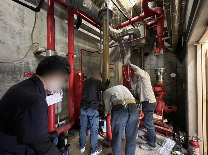 4人の作業員が新しい吸水管を垂直に支えながら、床の貫通部から消火水槽へ降ろしているところ