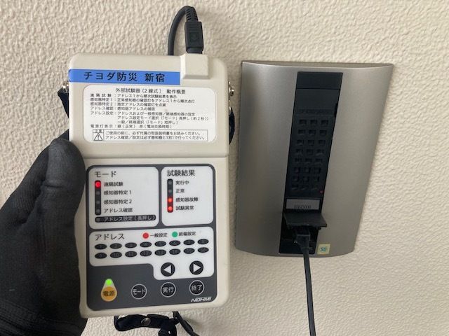 住戸外の壁に設置された遠隔試験機能付き感知器の子機に外部試験器を接続している様子